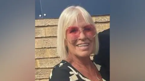 Janet Finch, a blonde woman with shoulder length hair and a fringe.
She is wearing a black dress with white floral designs and pink tinted sunglasses. Ms Finch is smiling at the camera.