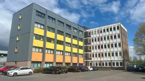 The car park at the back of Orchard House, showing two parts of the building, one of which is more modern, with grey, yellow and dark grey panels.