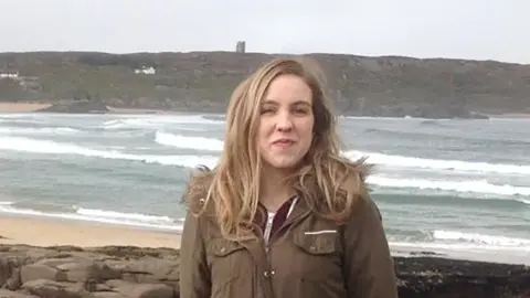 Natalie McNally. She has long blonde hair, wearing a long khaki coat and standing on a beach.