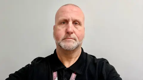 A man with very short hair and white stubble is standing in front of a blank wall, wearing a black shirt.