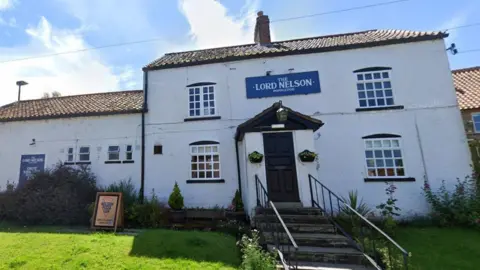 A large white building is pictured, with four windows and a protruding porch with a its own small triangular roof. There's hanging baskets on either side of the porch door, a light on the roof, and a blue sign saying 'The Lord Nelson Poppleton' above it. To the left there's an extension of the building, with four smaller windows and another blue The Lord Nelson Poppleton sign. There's stairs leading up to the door with hand railings on either side, and a green lawn at the front.