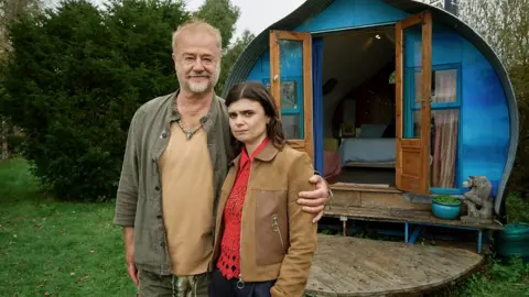 BBC/ BBC Studios Janie and her father, played by Owen Teale, stand outside a bright blue, wooden cabin with open wooden doors. The father has his arm around Janey's shoulder and she has an uncomfortable expression on her face. 