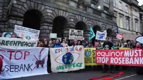 PA Media A group of protesters from climate organisations outside the Court of Session in Edinburgh. From left to right, one group are holding a greenpeace banner, which has the name of the organisation in green on a white banner. Below that is the words "Stop Rosebank" with stop in red and Rosebank in black. An image of an oil platform with a red X over the top is next to it. The next banner reads "Edinburgh" in multi-coloured letters and Greenpeace in green on a light-coloured banner either side of a globe in blue and green with a red eye shape surrounding it. The final banner is red with the words "equinor must stop Rosebank" in white letters.