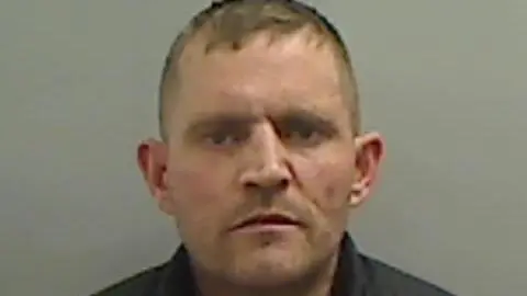 Mugshot of Dobie. He is clean shaven with short fair hair and has a menacing expression on his face.