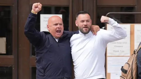 PA Media Two men with their arms around each others shoulders with their hands raised smile outside a court building
