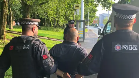 Two police officers in black uniform hold the arms of a man wearing handcuffs as they guide him towards a police van parked at a roadside.