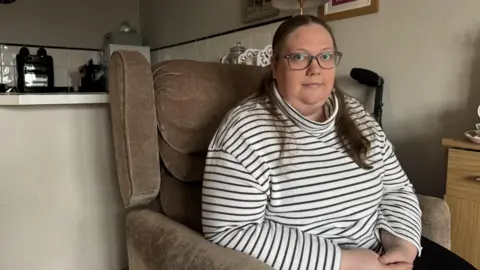 Hannah Roe/BBC A woman in a striped jumper sits in a modified chair facing the camera. 