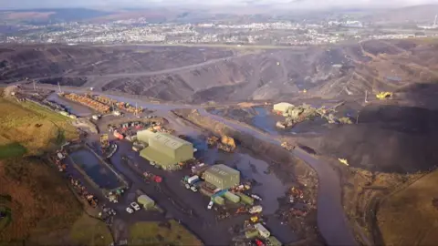 An aerial photograph of Ffos-y-Fran mine when it was operational. There is a large green building in the middle of the site with vehicles surrounding it and a larger crater of grey earth behind.