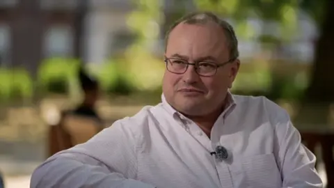 Middle aged man with glasses and a white shirt sitting on a park bench.