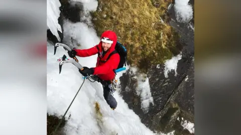 James Gibson Matt Le Voi ice climbing on a mountain. He is smiling up at the camera and is wearinga red jacket, helmet and rucksack, as well as using ice climbing equipment.