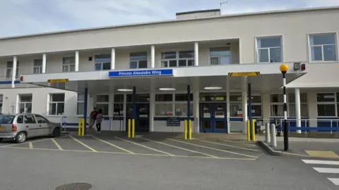 Royal Cornwall Hospital frontage with PRINCESS ALEXANDRA WING written above the main door on a blue sign. There are two people stood outside the two storey white building speaking. 