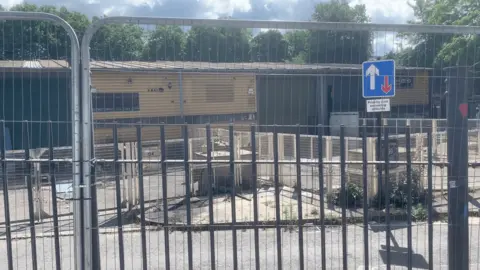 Kris Jalowiecki Temporary fencing in front of a collection of garages. There is a road in the foreground. 
