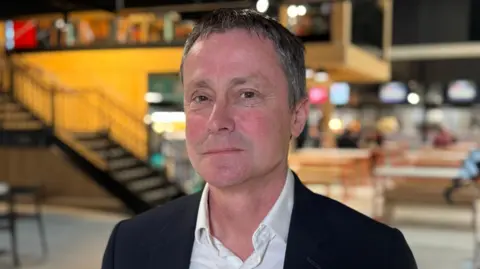 Andy Williams is wearing a dark blue suit and open-necked white shirt. He is looking at an angle into the camera and behind him is a colourful backdrop of a restaurant area in an open plan building, with yellow walls, large tables, food serving counters and a metal staircase leading up to a mezzanine.