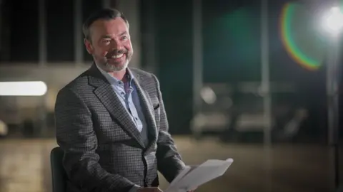 A man in a tweed suit jacker smiles