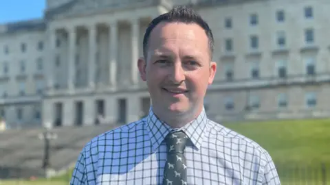 A man, with short black hair, wearing a white shirt with green squared lines on it, and a dark green tie. He is standing in front of the Stormont building. 
