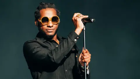 Leon Bridges is seen wearing yellow sunglasses and holding a microphone