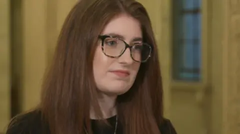 Connie Egan has long brown hair and is wearing black-rimmed glasses.
She is wearing a black top and necklace while standing in Stormont.