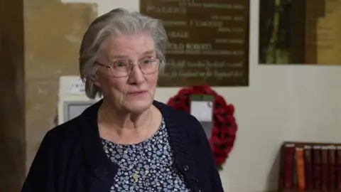 Alison Hoddell stands in the church with the plaque and a poppy wreath behind her. She has short grey hair and is wearing a blue patterned dress and navy cardigan. 