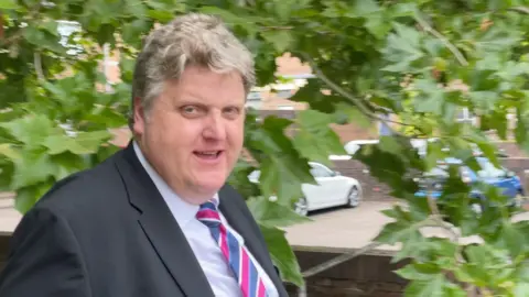 BBC Gregory Hill wearing a suit and multi-coloured smile, with a tree behind him