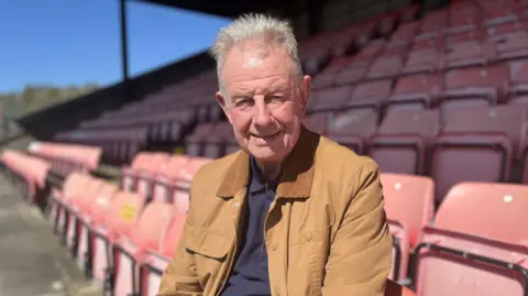 Man in a brown jacket and navy shirt sits in sunlit stadium seating, smiling at the camera, with rows of empty red seats stretching behind him.