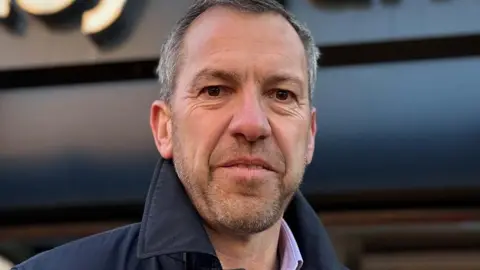 BBC/Paul O'Gorman Head shot of a smiling Alistair Bradley. He has short grey hair and stubble