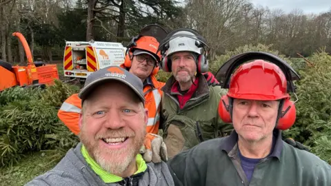 Four men, three of them in work helmets and headphone and one man wearing a grey cap, smiling for the camera while taking a selfie. You can see Christmas trees being shredded in the background.