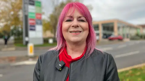 A woman with pink, shoulder-length hair, wearing a black leather jacket and a red top underneath