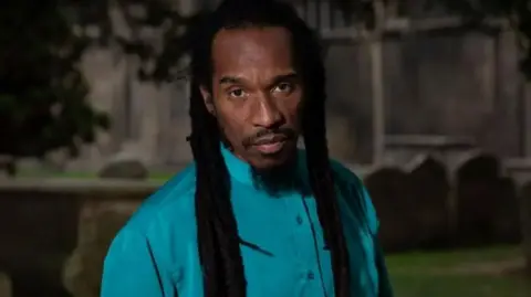 A man with dreadlocks and a blue shirt looks into the camera.