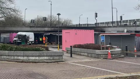 Tony Fisher/BBC Some market stalls which have clearly suffered fire damage under a road bridge. There are other market stalls with their shutters down. A white van and a pink container can be seen behind some bollards and a small hedge in a concrete bed.