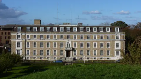 Shire Hall is four-storeys high Georgian building and has 14 large windows on each level. There is a garden to its front, and a blue car is parked outside the front entrace. 