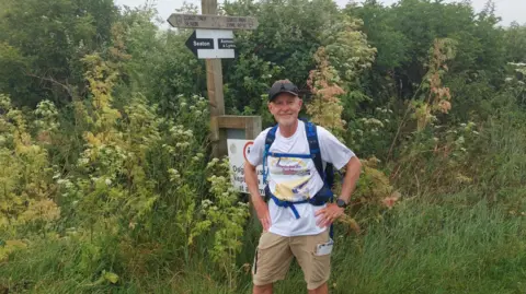 Handout Brown in beige cargo shorts and a white t-shirt with a motif on it. He has a blue rucksack on his back and is wearing a khaki cap and sunglasses while smiling at the camera with his hands on his hips. He is standing in front of greenery with signs for Seaton and Lyme Regis next to him. 