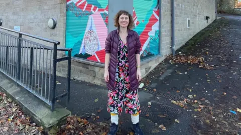 Roland Sebestyen A woman is standing in front of a building smiling at the camera.  She is wearing a long purple coat with a bright black pink and red flowery dress underneath with white leg warmers and black trainers.