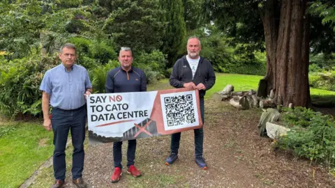 Three men stand on a garden path surrounded by green plants with a banner that reads say no to the Cato Data Centre