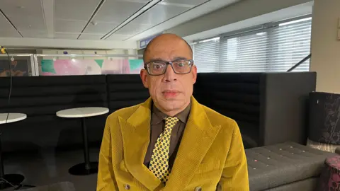 A bald man wearing dark glasses sits in a chair and looks at the camera with a straight face. He wears a brown shirt, yellow and brown tie, and yellow cuordroy jacket.