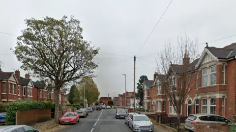 Stroud Road in Gloucester. There are red brick houses on both sides and cars parked on the side of the road. The sky is grey.