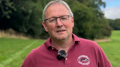 A man with short grey hair, wearing black metal rimmed glasses, and a red polo shirt. He is pictured standing against greenery in a field.
