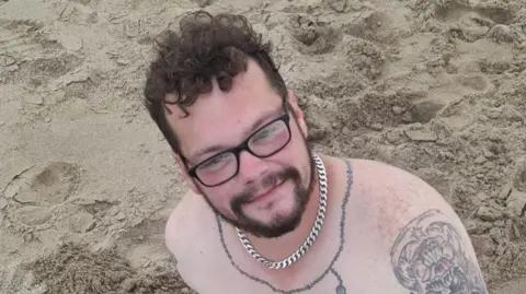 Family handout A picture of Aaron Keightley smiling as he looked up towards the camera. He is sitting on a beach, shirtless and wearing a silver chain. Tattoos can be seen on his chest and upper arm.