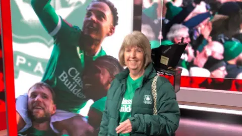 Brian Cardell Plymouth Argyle fan Julie Cardell stood in the BBC Spotlight studio wearing her Argyle kit and coat. She is stood in front of a screen with Plymouth Argyle players pictured in it.