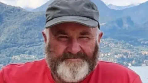 A man with a brown and grey beard is looking into the camera. He is wearing a red t-shirt and grey baseball cap. In the background we can see a body of water and mountains. 