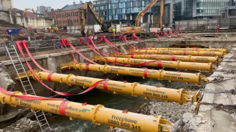 Simon Thake/BBC A construction site with several yellow pipes stretched across an uncovered river with rubble scattered about on both sides.