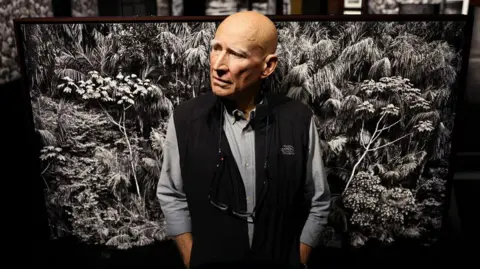 Sebastião Salgado standing in front of a black and white photo of trees in the Amazon