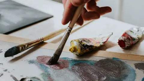 Getty Images A close-up of a right hand holding a paintbrush as it daubs red oil or acrylic paint onto a canvas which has blue swirls of colour on it. There is another paintbrush resting on the canvas and two open tubes of paint as well, one resting on the canvas and the other resting on a table.