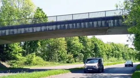 A white van travels underneath one of the bridges. A car is also approaching the spot. The bridge has metal railings running along the top and there are trees to the sides of the road.