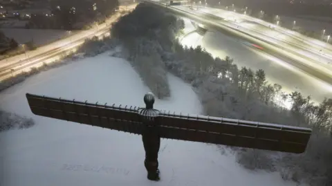 Snow surrounds the Angel of the North. The roads surrounding it are lit by streetlights