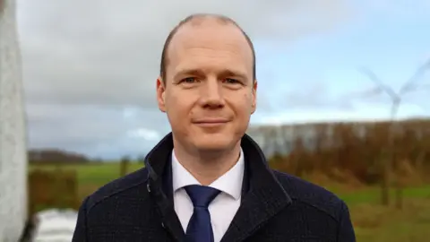 Gordon Lyons, who is mostly bald, smiles at the camera. He is wearing a dark coat over a white shirt and navy tie. Behind him is Irish countryside; grass and brush hedges. 
