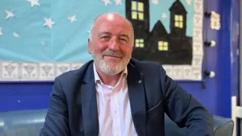 Kurt Roosen, who is wearing a navy blue suit jacket with a white shirt. He has short grey hair and a grey beard. He sits in front of a colourful display.