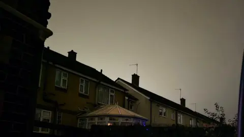 Helen Attridge A glow in the night sky can be seen, in the foreground there are terraced houses