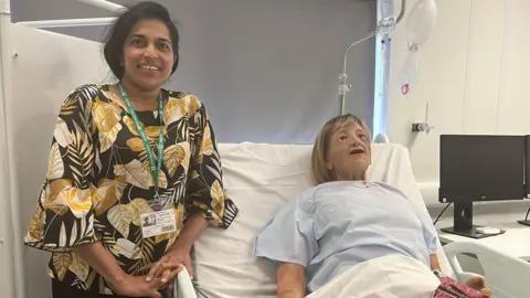 Senior care worker Sajee Gunarathna stands next to the bedside of a model patient called 'Dorris'. Sajee is wearing a black and yellow floral top and has long black hair. 
