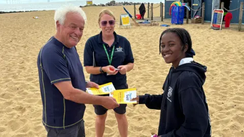 Jodie Halford/BBC A man presents a school pupil with a leaflet and a badge for having completed a Swim Safe course. A woman wearing a lanyard and sunglasses stands in the background.