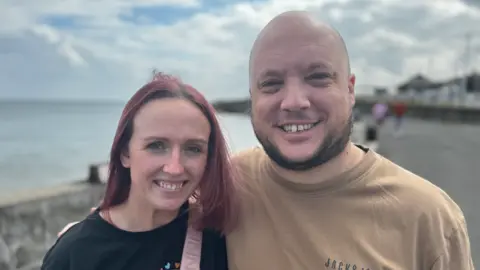 The BBC's Kayley Avery, with dyed red hair and a black T-shirt, stands next to her husband David, who is wearing a brown T-shirt.